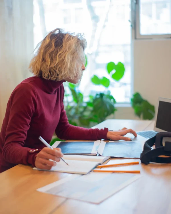 Frau prüft am Schreibtisch Unterlagen und macht sich Notizen neben einem Laptop