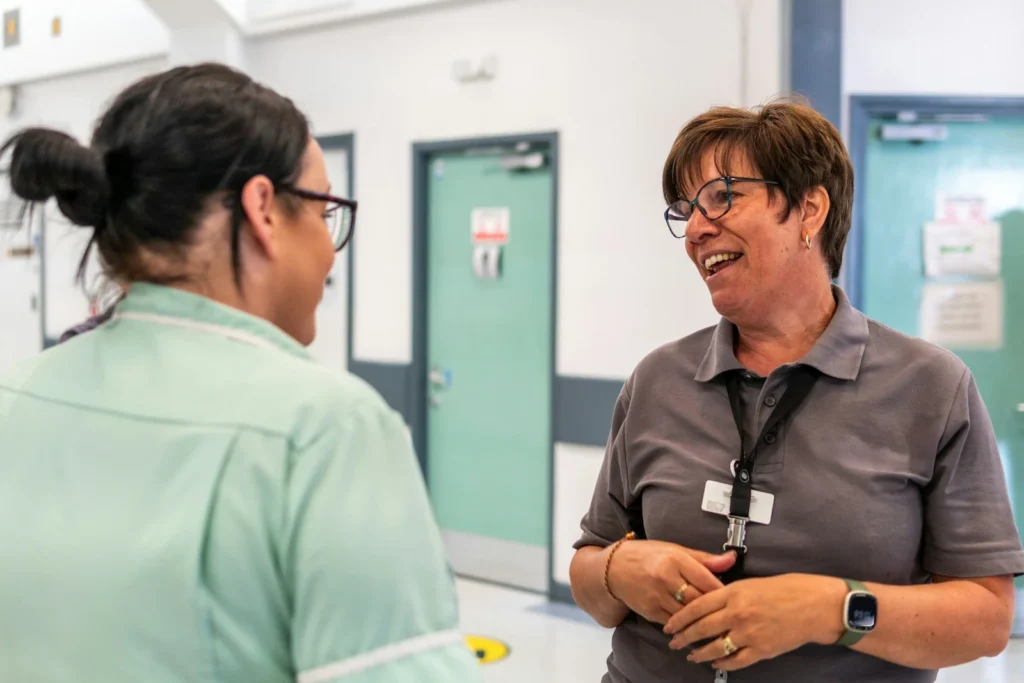 Zwei Menschen unterhalten sich lachend auf einem Klinikflur – eine Person in mintgrüner Dienstkleidung, die andere im grauen Poloshirt mit Namensschild